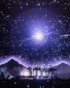 Two musicians perform on stage with a starry night sky and mountain backdrop, illuminated by bright lights and visual effects.